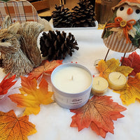 A beige candle with a pumpkin pie scent, displayed on a white surface surrounded by autumn-themed decorations including leaves, a squirrel, and a snowman.
