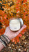 Thankful jar candle with a white substance inside, in front of a background with autumn-colored leaves.