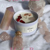 A soy candle with a blend of aromatic herbs, warm amber, and florals, topped with dried rose petals, Himalayan pink salt, and a Rose Quartz heart, presented in a white jar surrounded by decorative stones and a small glass bottle.